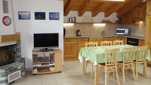 A cozy kitchen with wooden cabinets, a dining table set for four, a flat-screen TV, and a stone fireplace. - Hilltop Apartment With Views (Veysonnaz)