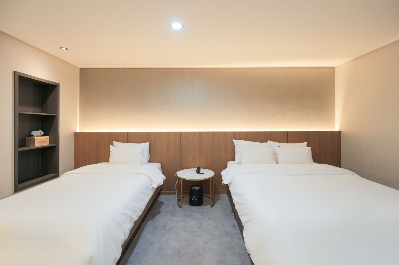 Hotellihuone, jossa on kaksi erillistä sänkyä, pieni pyöreä pöytä, kirjahylly ja kattovalaisin.