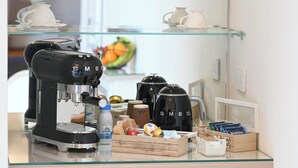 A coffee station with a Smeg espresso machine, a glass display case with teapots, and a wooden box with snacks and a water bottle. - Dimora Bianca Luxury (Conversano)