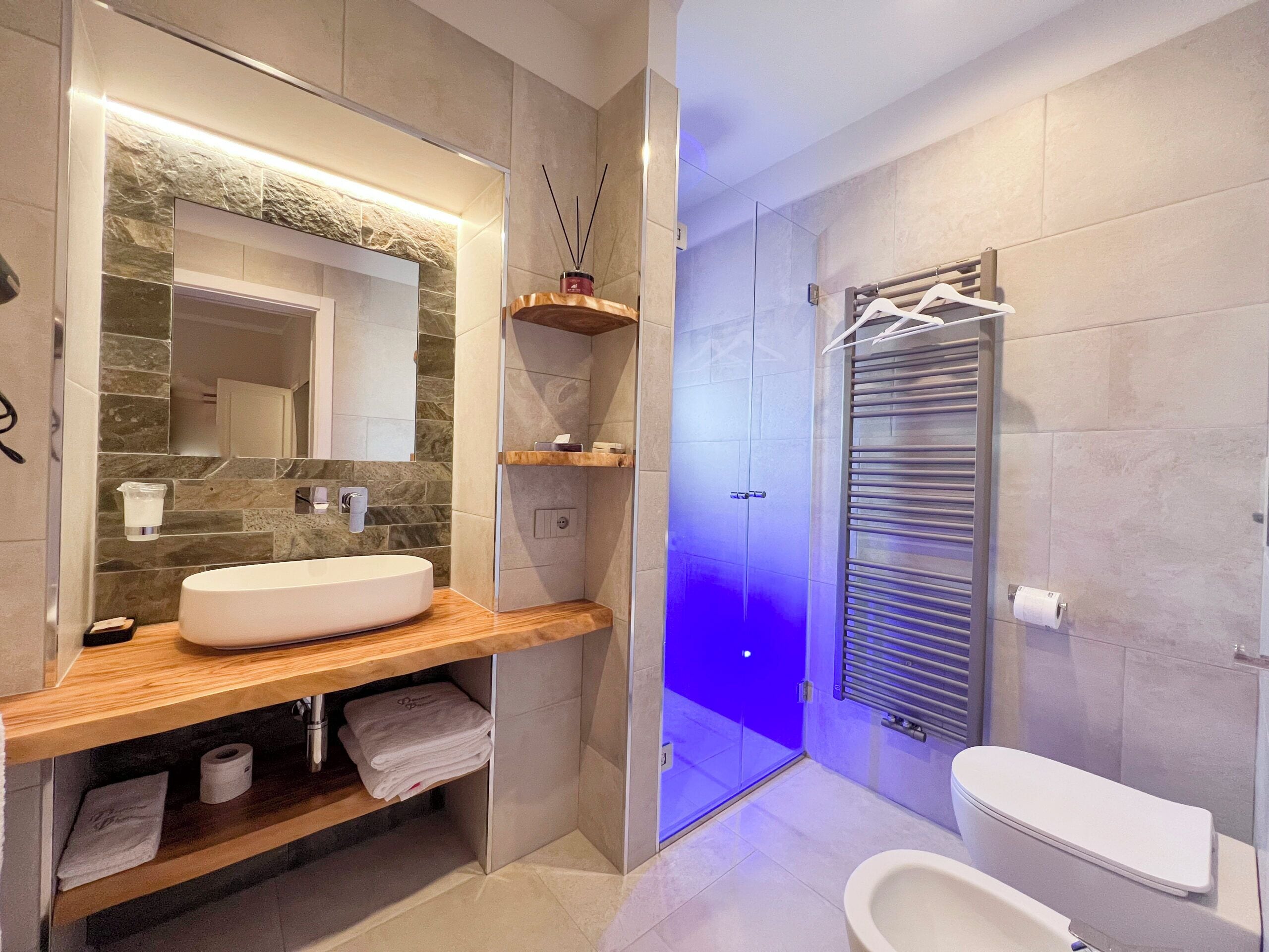 A modern bathroom with a large mirror, a white sink, a shower with blue lighting, and a toilet.