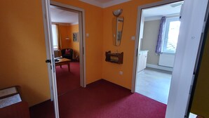 A hallway with a red carpet leading to a room with a bed and a window. There is a door on the left and a window on the right.