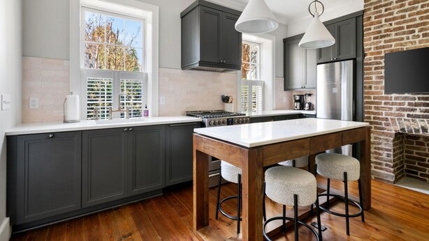 A modern kitchen with dark cabinets, a central island with stools, a brick accent wall, and a large window. - Thomas Waring House | Unit B | Sleeps 4 (Charleston)