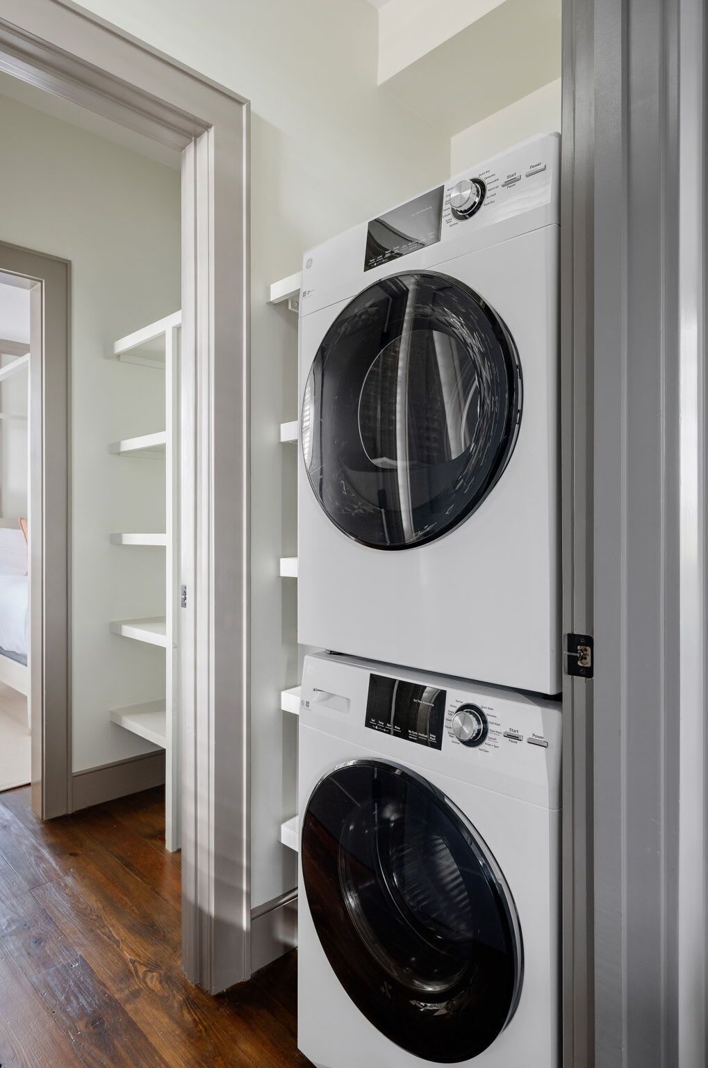 A laundry room with two stacked front-loading washing machines and a white shelving unit.