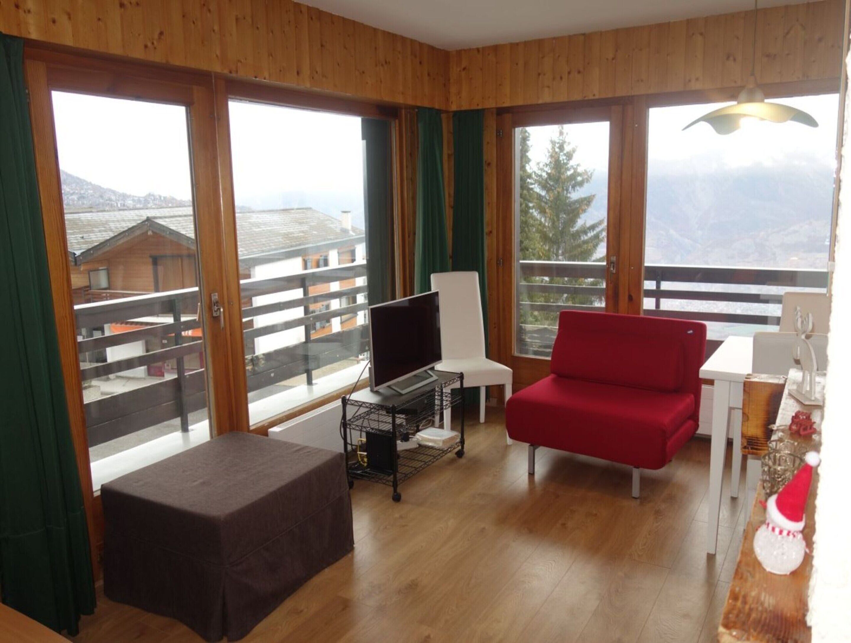 Une pièce au plancher de bois, avec un canapé rouge, un téléviseur posé sur un meuble et un balcon offrant une vue sur les bâtiments et les montagnes.