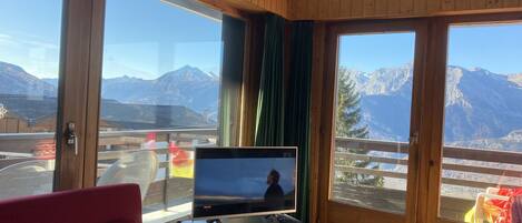 A room with a TV on a stand, a red sofa, and a view of mountains through large windows.
