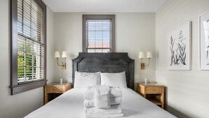 Una cama bien hecha con sábanas blancas y una pila de toallas blancas, flanqueada por dos mesitas de noche con lámparas, en una habitación con un ventanal grande e impresión botánica enmarcada en la pared.