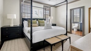 A four-poster bed with a canopy, a bedside table with a lamp, and a view of the bathroom through an open door. - Rosemary House - C | Perfect for Weddings + Events (Charleston)
