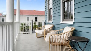 A porch with wicker chairs and a small table, a white railing, and a blue building in the background. - Rosemary House - C | Perfect for Weddings + Events (Charleston)