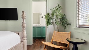 Un dormitorio con cama, una silla, una mesita, televisión y vista al baño.