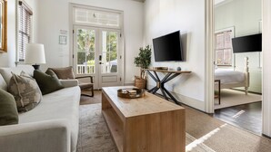 A well-lit living room with a white sofa, a wooden coffee table, and a TV on a black stand. There is a door leading to a balcony with a view of greenery. - Harleston House, Unit B, Sleeps 6 (Charleston)