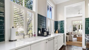 A modern kitchen with white cabinetry, a large window with blinds, a coffee maker, and a view to the living area. - Harleston House, Unit B, Sleeps 6 (Charleston)