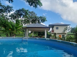 Una piscina con borde de azulejos, un pabellón cubierto y una casa de dos pisos con balcón.
