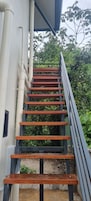 Una escalera de madera con barandillas negras que conduce a un edificio con paredes exteriores blancas y tuberías visibles.