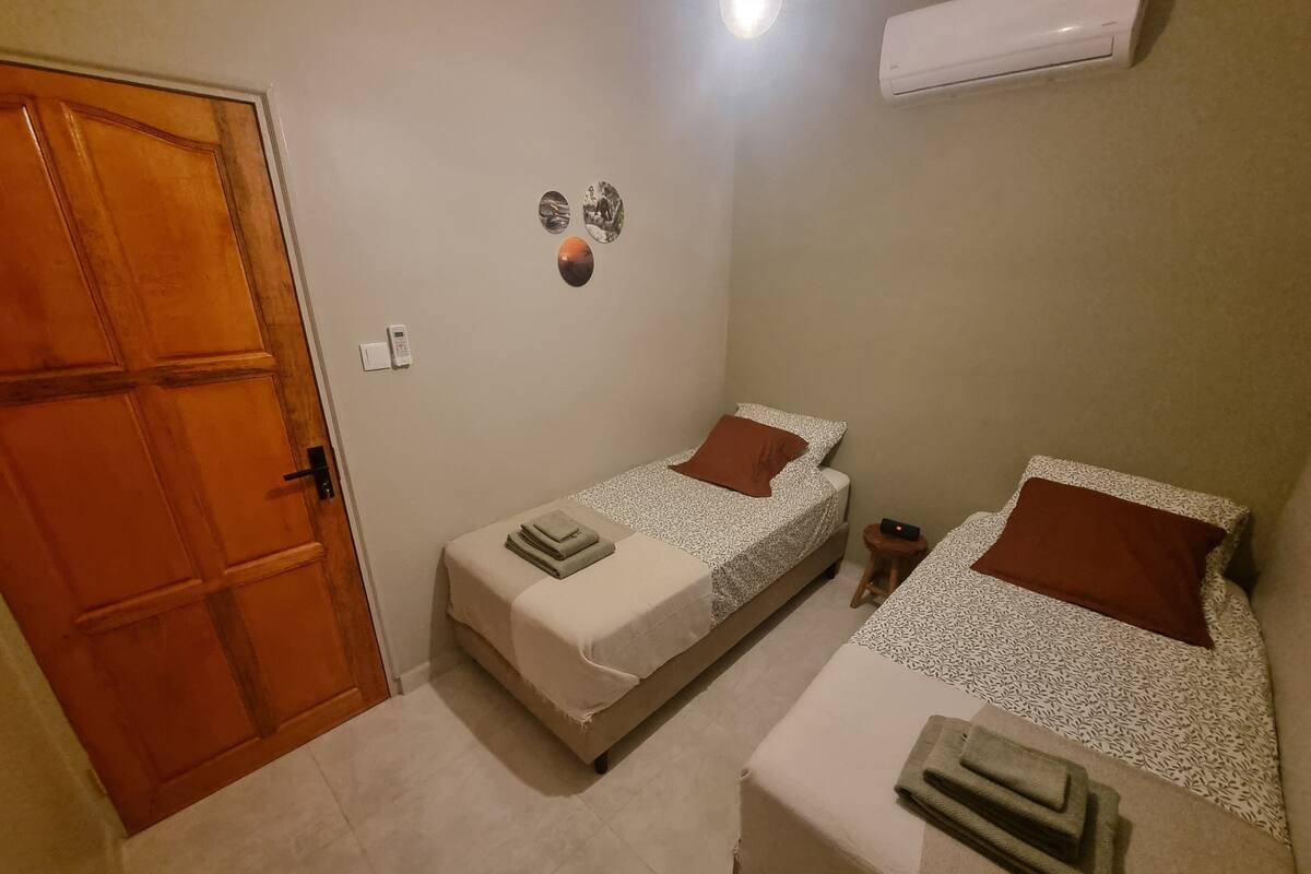 Ein kleines, sauberes Zimmer mit zwei Betten, einer Holztür, einer Wand-Klimaanlage und einem kleinen Tisch mit Telefon.