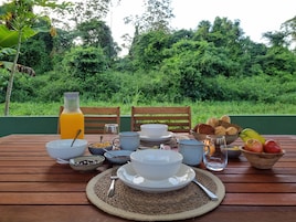 Un desayuno servido en una mesa de madera al aire libre, con diferentes platos, una botella de jugo de naranja y un fondo de vegetación exuberante.