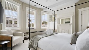 A bedroom with a four-poster bed, a sitting area with a chair and table, and large windows with blinds. - The Gardenia House | Sleeps 20 | Multi-Unit Rental (Charleston)