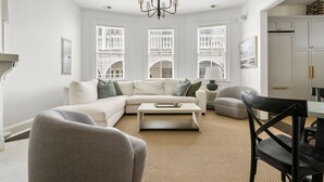 A modern living room with a white sofa, a coffee table, and a dining area with a table and chairs. - The Gardenia House | Sleeps 20 | Multi-Unit Rental (Charleston)