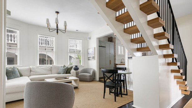 A modern living room with a white sofa, grey armchair, and a dining area with a table and chairs. A staircase with wooden steps leads to an upper level. - The Gardenia House | Sleeps 20 | Multi-Unit Rental (Charleston)