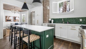 A modern kitchen with a central island, white cabinetry, green backsplash, and a brick accent wall. - The Gardenia House | Sleeps 20 | Multi-Unit Rental (Charleston)