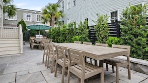 A patio with a wooden table and chairs, surrounded by greenery and a white building. - The Gardenia House | Unit A | Sleeps 7 (Charleston)