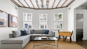A modern living room with a sectional sofa, a coffee table, and a wooden side table. There are three windows with blinds, a chandelier, and a refrigerator in the background. - The Gardenia House | Unit A | Sleeps 7 (Charleston)