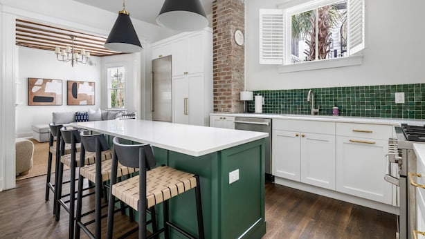 A modern kitchen with a central island, white cabinetry, green backsplash, and a brick accent wall. - The Gardenia House | Unit A | Sleeps 7 (Charleston)
