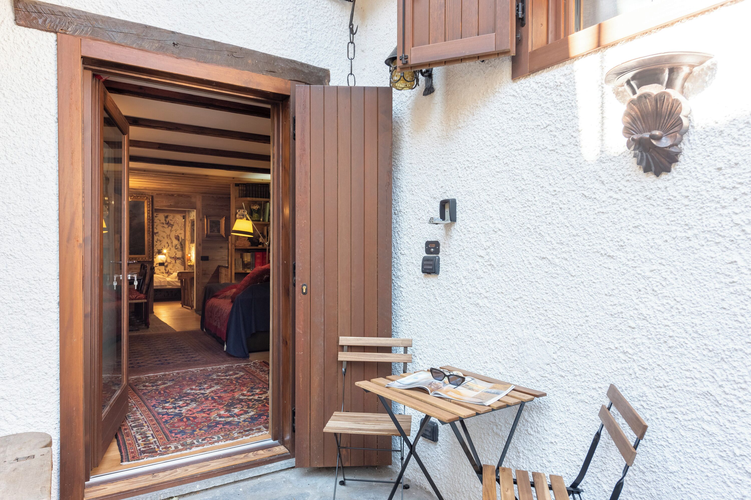 Une porte en bois ouverte donne sur une pièce où se trouve un tapis à motifs, une petite table avec une chaise et un luminaire fixé au mur.