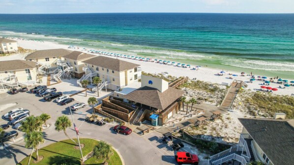 An aerial view of a beachfront hotel with multiple buildings, a parking lot, and a beach with umbrellas.