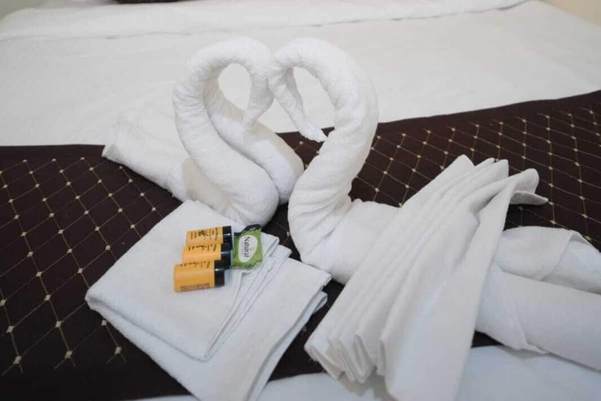 Un lit bien fait, avec du linge de lit blanc, deux oreillers marron et une serviette pliée en forme de cœur posée sur une couverture à motifs foncés.