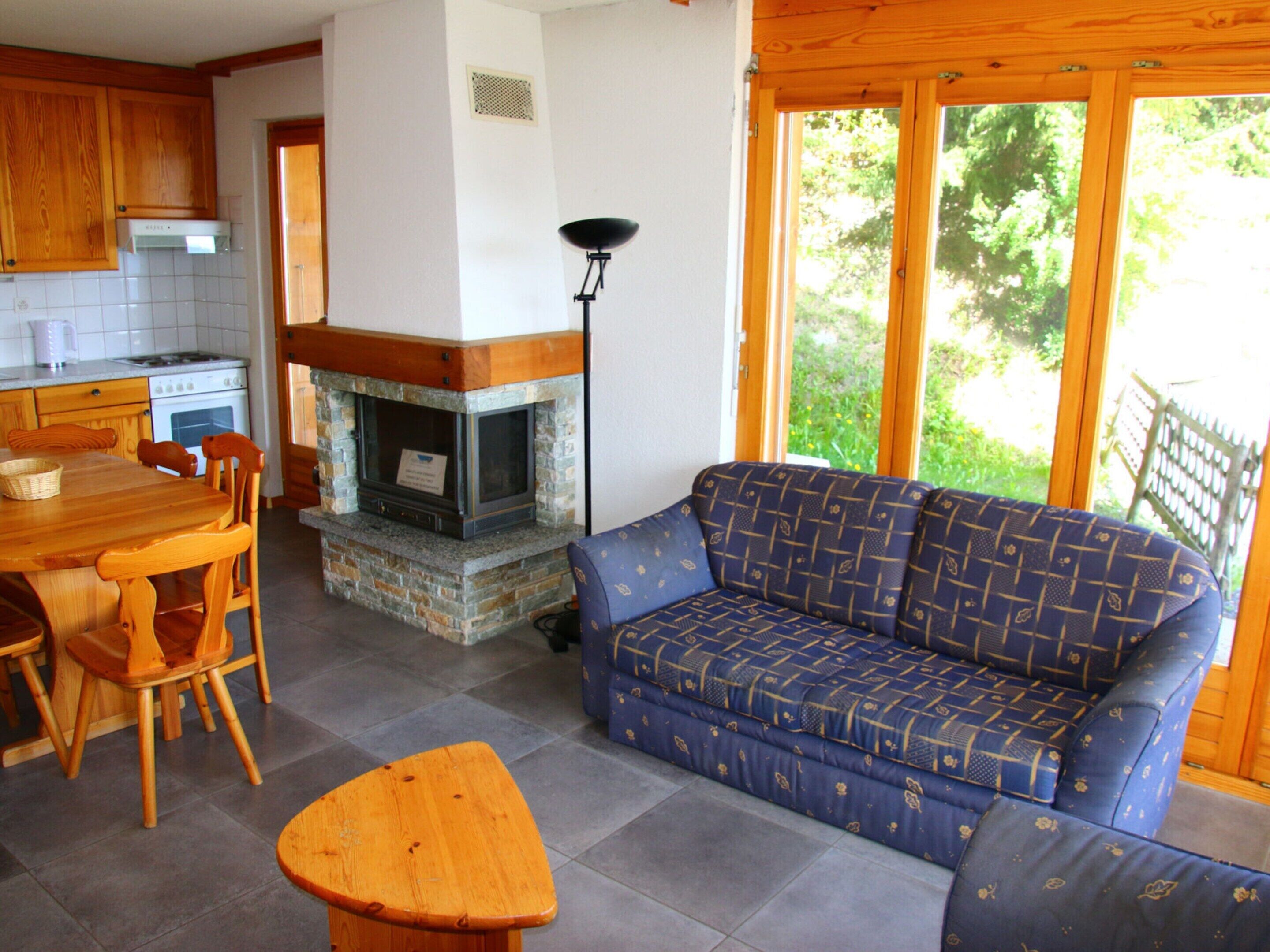 A cozy living area with a stone fireplace, a blue patterned sofa, wooden furniture, and a view of greenery through large windows.