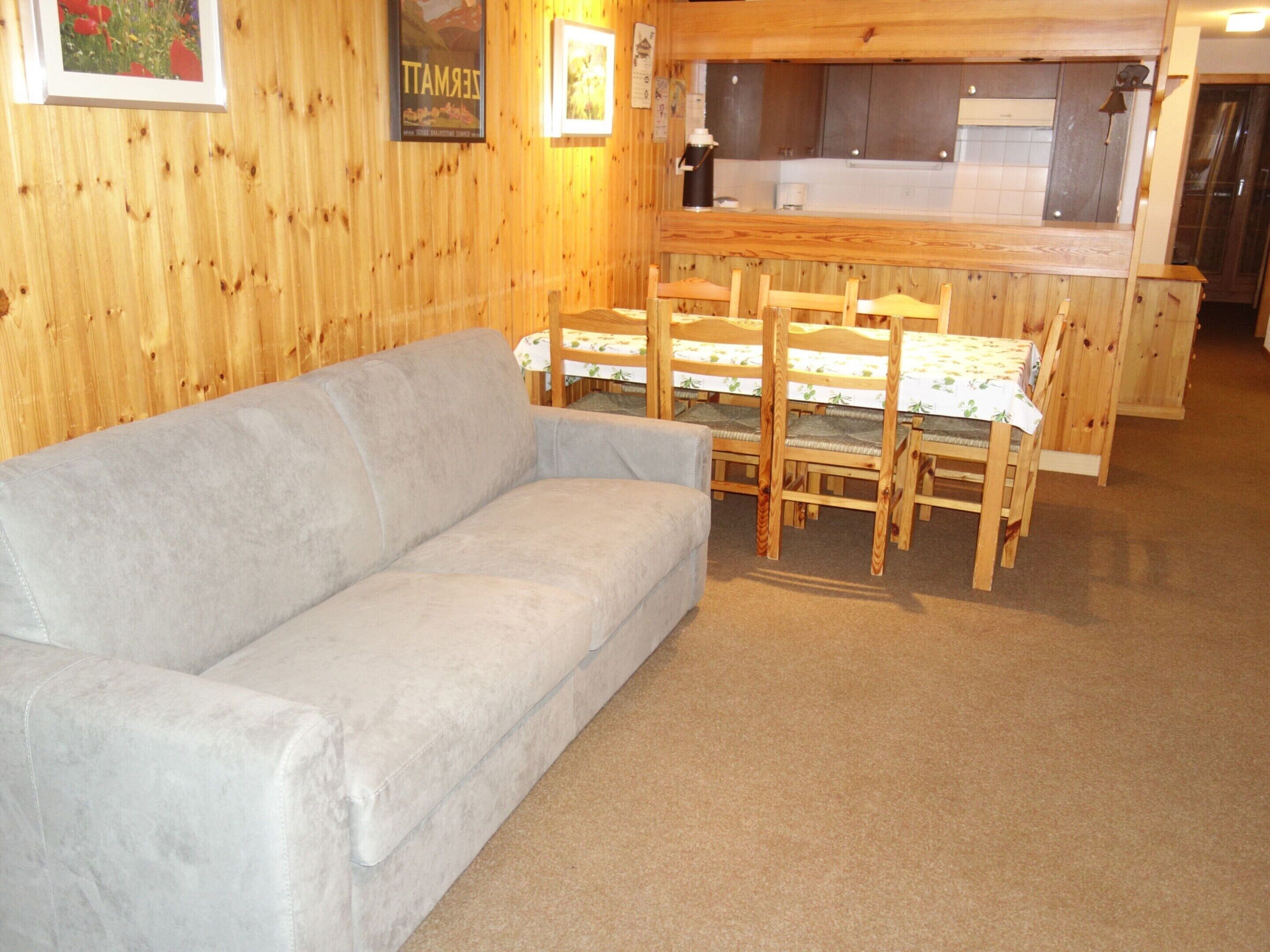 Ein gemütliches Zimmer mit grauem Sofa, einem Holzespeisetisch und Stühlen. Es gibt eine Küchenzeile mit Schränken und einem Spülbecken.