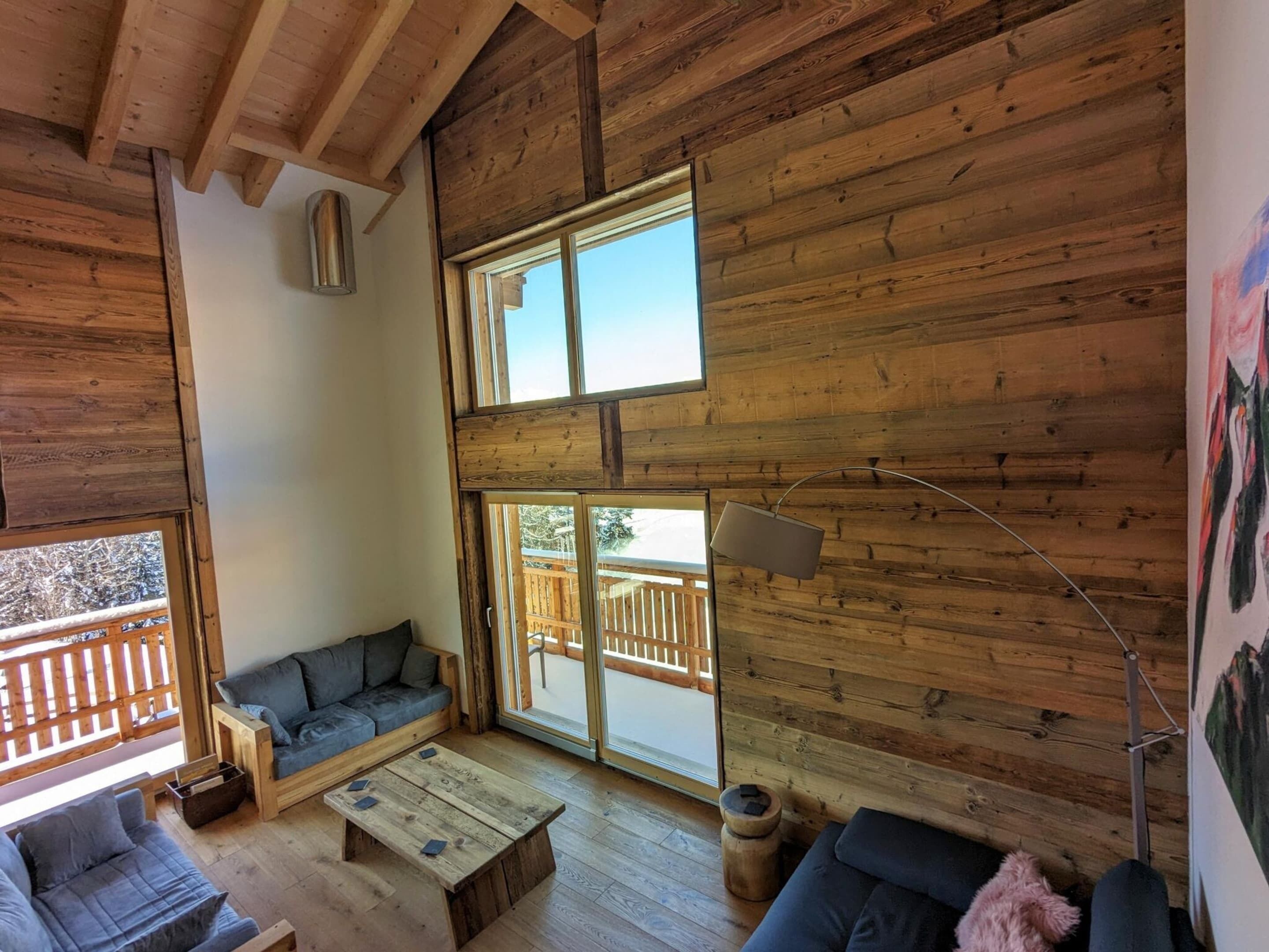 A cozy cabin interior with wooden walls, a sofa, a coffee table, and a large window offering a view of a wooden deck.