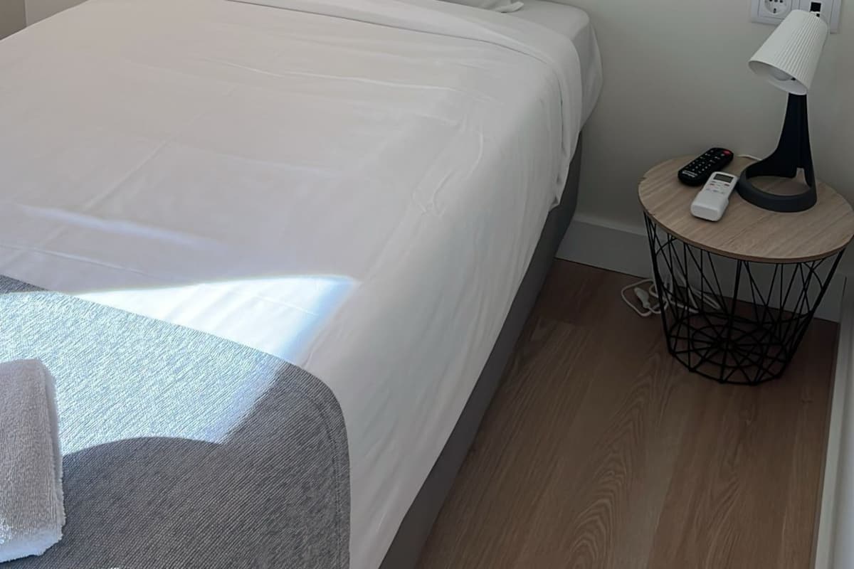 A single bed with white bedding, a bedside table with a lamp, and a power outlet on the wall.