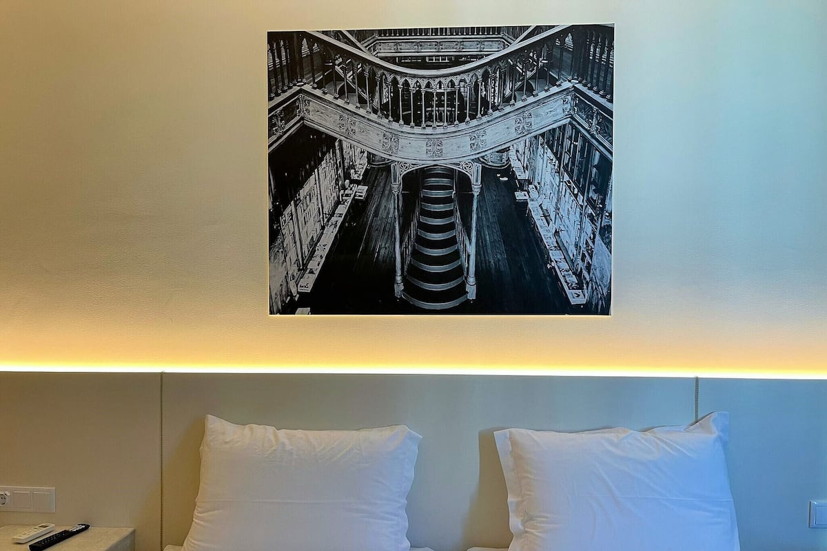 A hotel room with a bed, two pillows, a nightstand with a remote control, and a framed picture of a staircase.