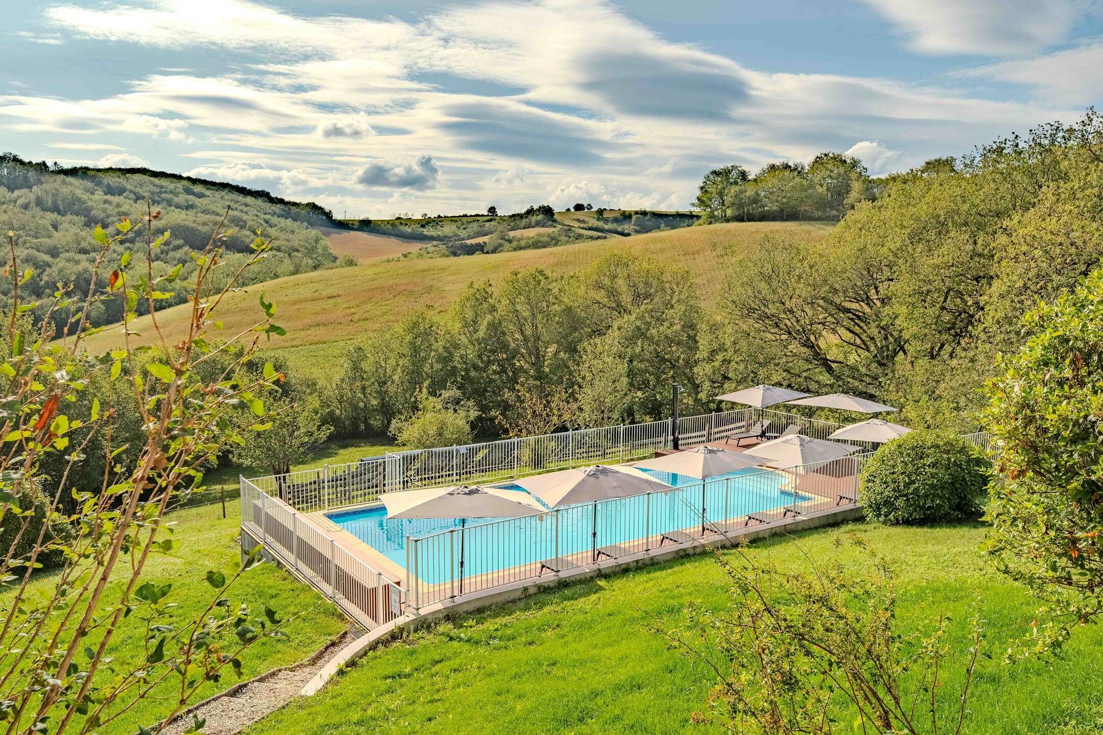 Ein Schwimmbad mit umzäuntem Deck und Sonnenschirmen, umgeben von üppiger Vegetation und Hügeln.