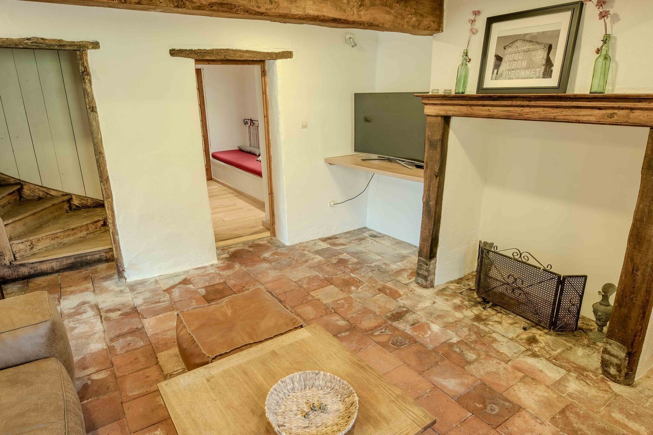 Une pièce rustique avec un plancher en bois, un lit recouvert d’une literie rouge, un téléviseur posé sur une étagère en bois et une cheminée dotée d’un paravent en métal.