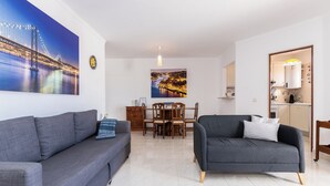 Uma sala de estar moderna com dois sofás cinzentos, um espaço de refeições com uma mesa de madeira e cadeiras, e uma cozinha visível através de uma porta aberta.