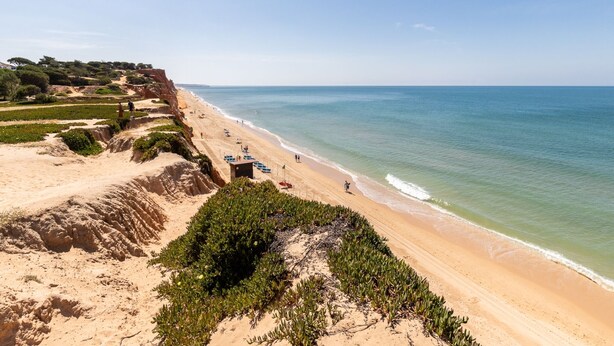 Paisagem costeira com praia arenosa, um penhasco com vegetação e céu claro.
