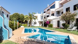 Uma piscina rodeada por um edifício branco com varandas, uma parede de azulejos e um céu claro.