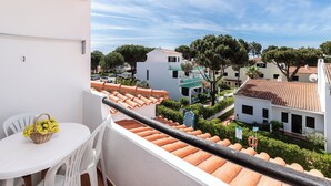 Uma varanda com mesa e cadeiras brancas, com vista para um pátio com edifícios e árvores.