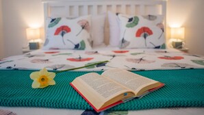 Un lit recouvert d’une couvre-lit à motifs floraux, une seule fleur jaune et un livre ouvert posés sur une couverture tricotée de couleur turquoise.