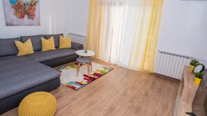 Un salon moderne avec un canapé d’angle gris, des coussins jaunes, un petit pouf rond jaune, une table basse blanche et un tapis coloré.