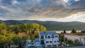 Une maison moderne à deux étages, dotée d’un toit plat, entourée d’une végétation luxuriante et avec une chaîne de montagnes en toile de fond.