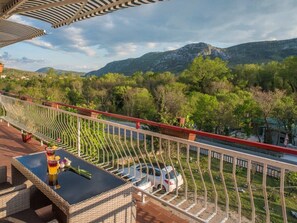 Un balcon avec vue sur une montagne, une voiture garée en contrebas et une table avec des boissons et des fleurs.