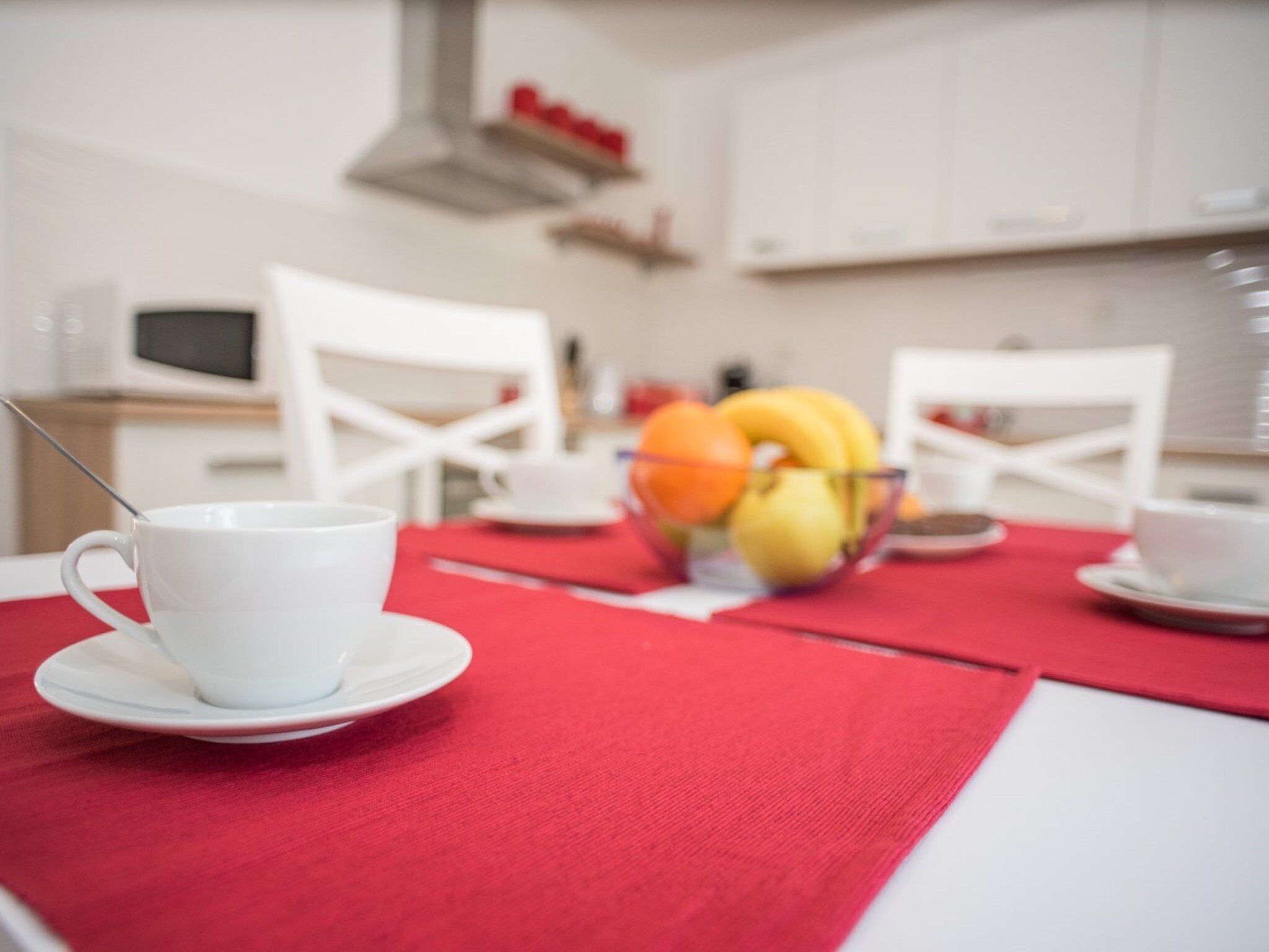 A dining table with a red tablecloth, white cups, a bowl of fruit, and a white chair.