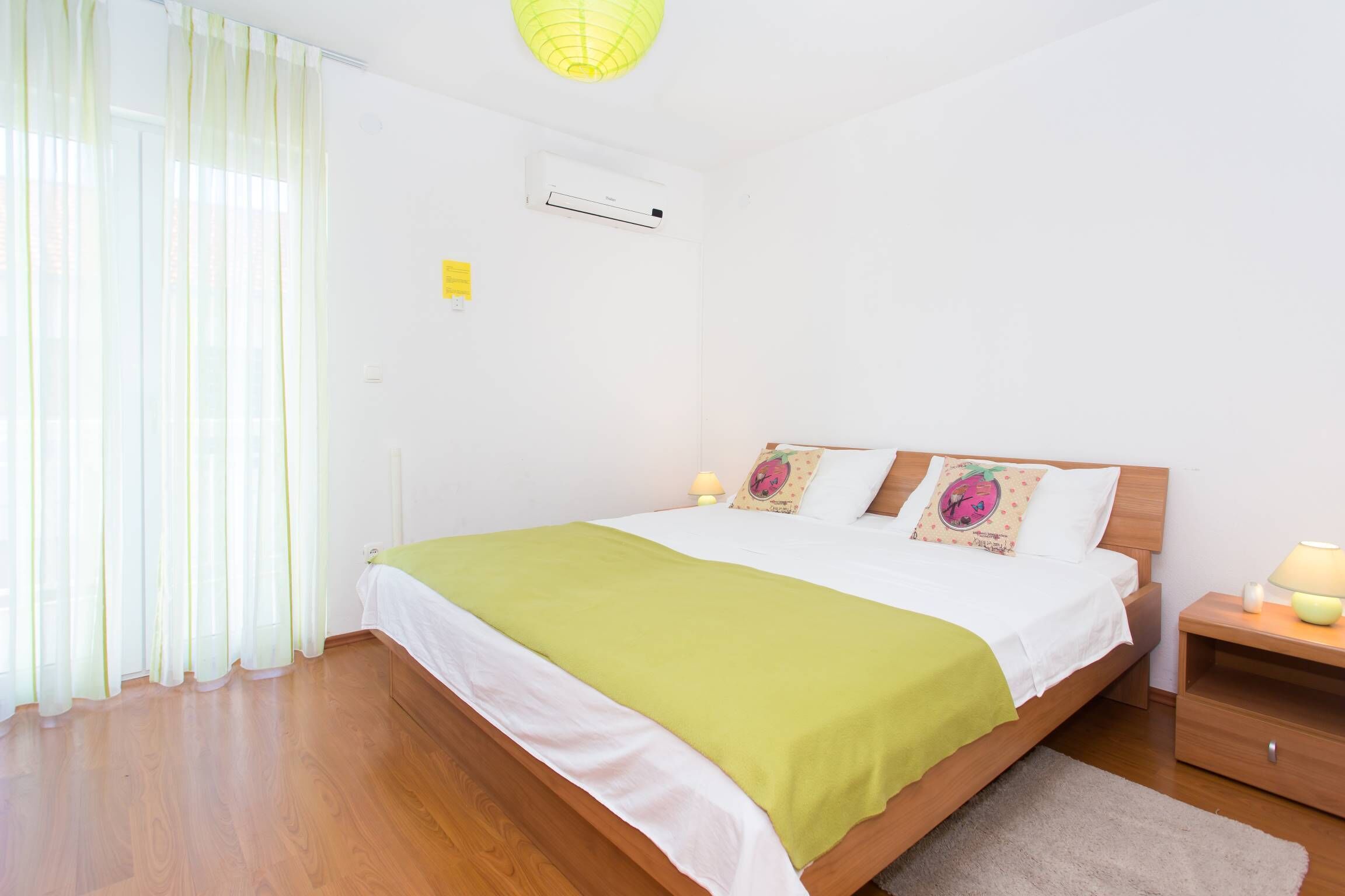 A bedroom with a wooden bed, white bedding, a green throw, a nightstand with a lamp, and a window with sheer curtains.