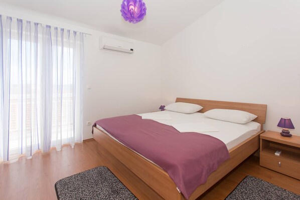 A bedroom with a wooden bed, white bedding, a purple lamp, and a window with sheer curtains. - Charming Apartments With Private Balcony and City View Seget Donji (Seget)