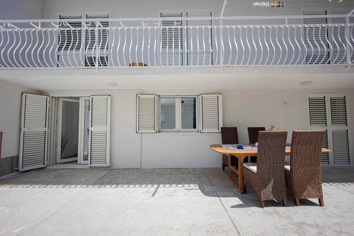 A covered patio with a table and chairs, white shutters on windows, and a white railing.