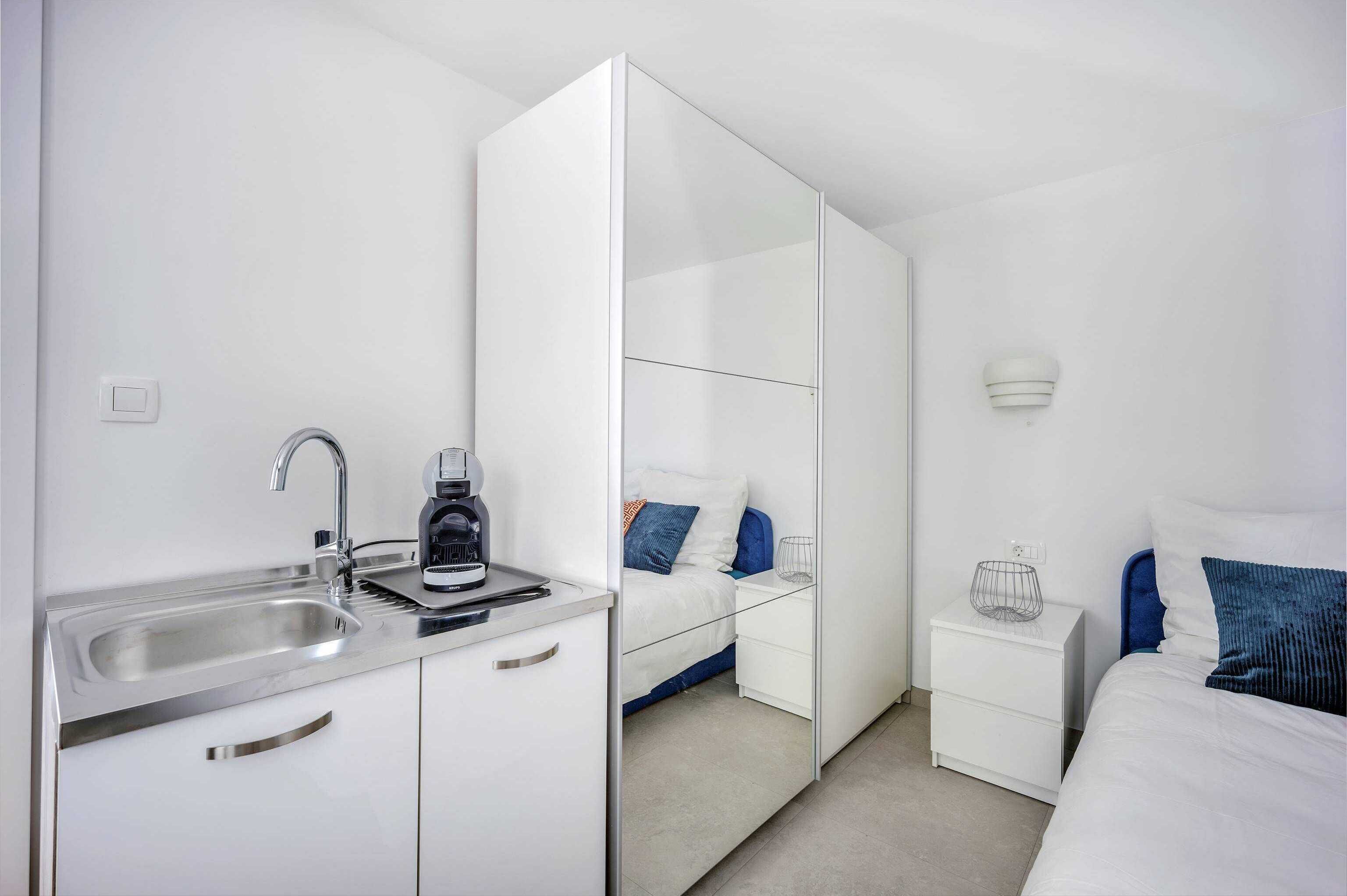 A modern hotel room with a kitchenette, a bed with blue and white bedding, a bedside table with a lamp, and a large mirror.