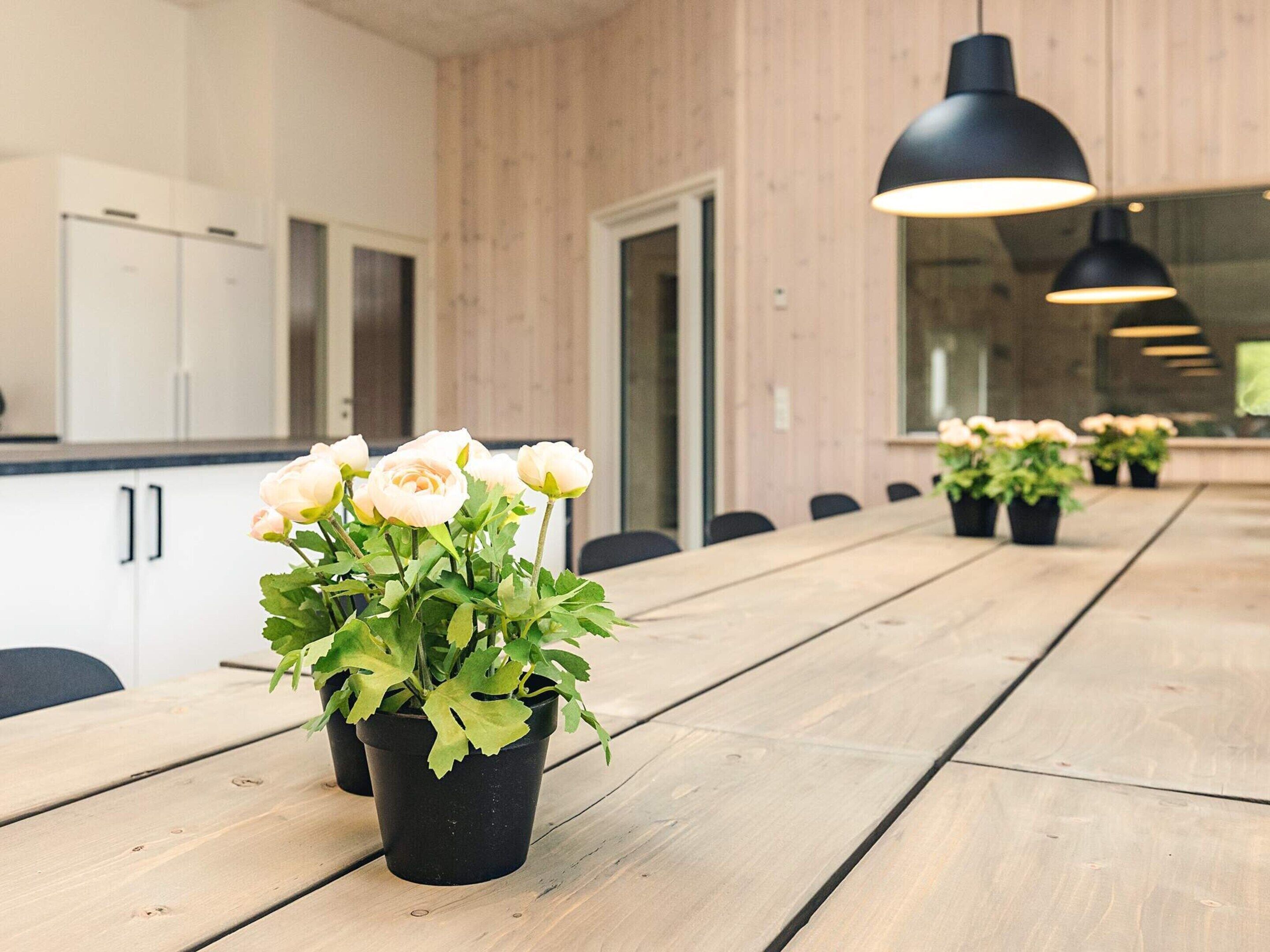 Une longue table en bois, ornée de pots de fleurs noirs, et en arrière-plan, une cuisine moderne.